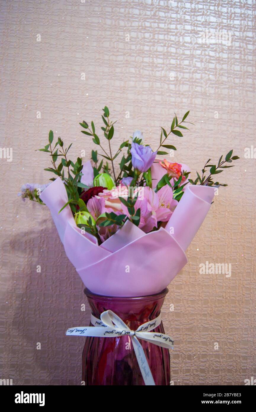 Ein zartes Blumenstrauß verschiedener Blumen in einem hellrosa Wrapper steht vor einem Hintergrund rosafarbener Tapeten in einer Hauseinstellung. Vertikales Foto. Stockfoto