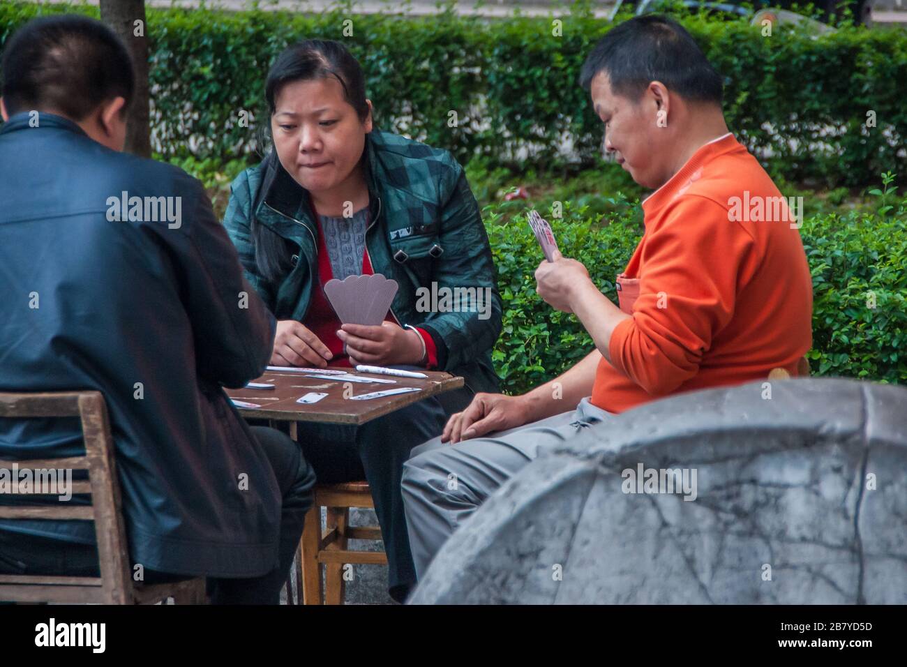 Guilin, China - 11. Mai 2010: Stadtzentrum. Auf Stühlen und Bank spielen Frau und 2 Männer chinesische Karten mit großem Fokus und Aufmerksamkeit. Grünes Laub im Rücken. Stockfoto
