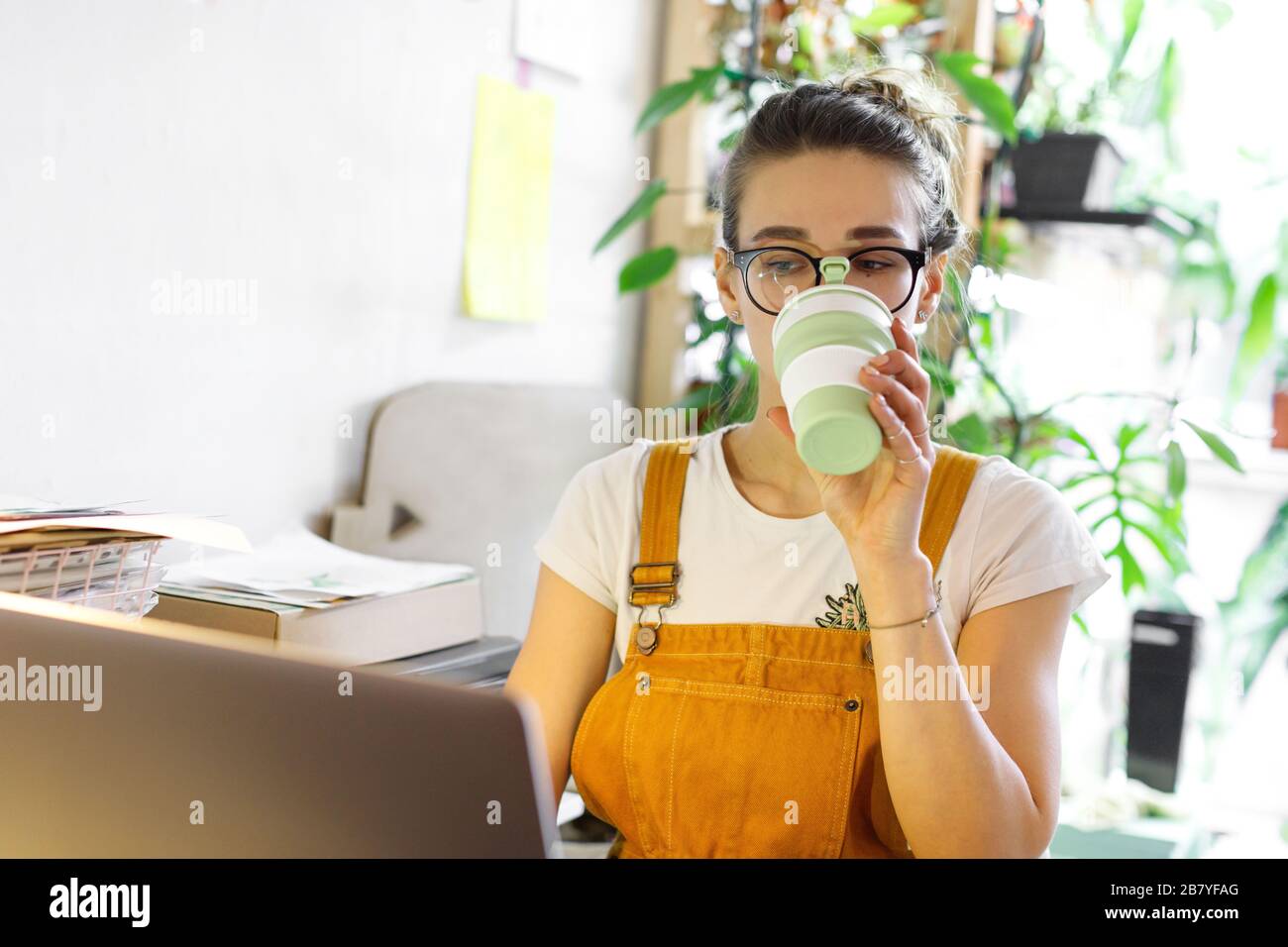 Junge Gärtnerin in Brille mit Laptop, kommuniziert im Internet mit Kunden im Garten/Gewächshaus und trinkt Kaffee/Tee aus wiederverwendbarem Becher Stockfoto