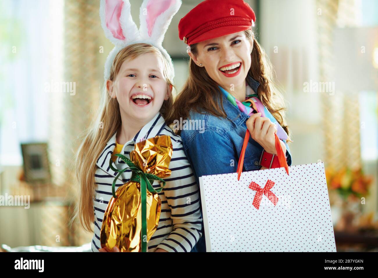 Portrait lächelnder eleganter Mutter und Tochter mit in Goldfolie gewickeltem großen osterei und Einkaufstaschen im modernen Wohnzimmer im sonnigen Frühling d Stockfoto