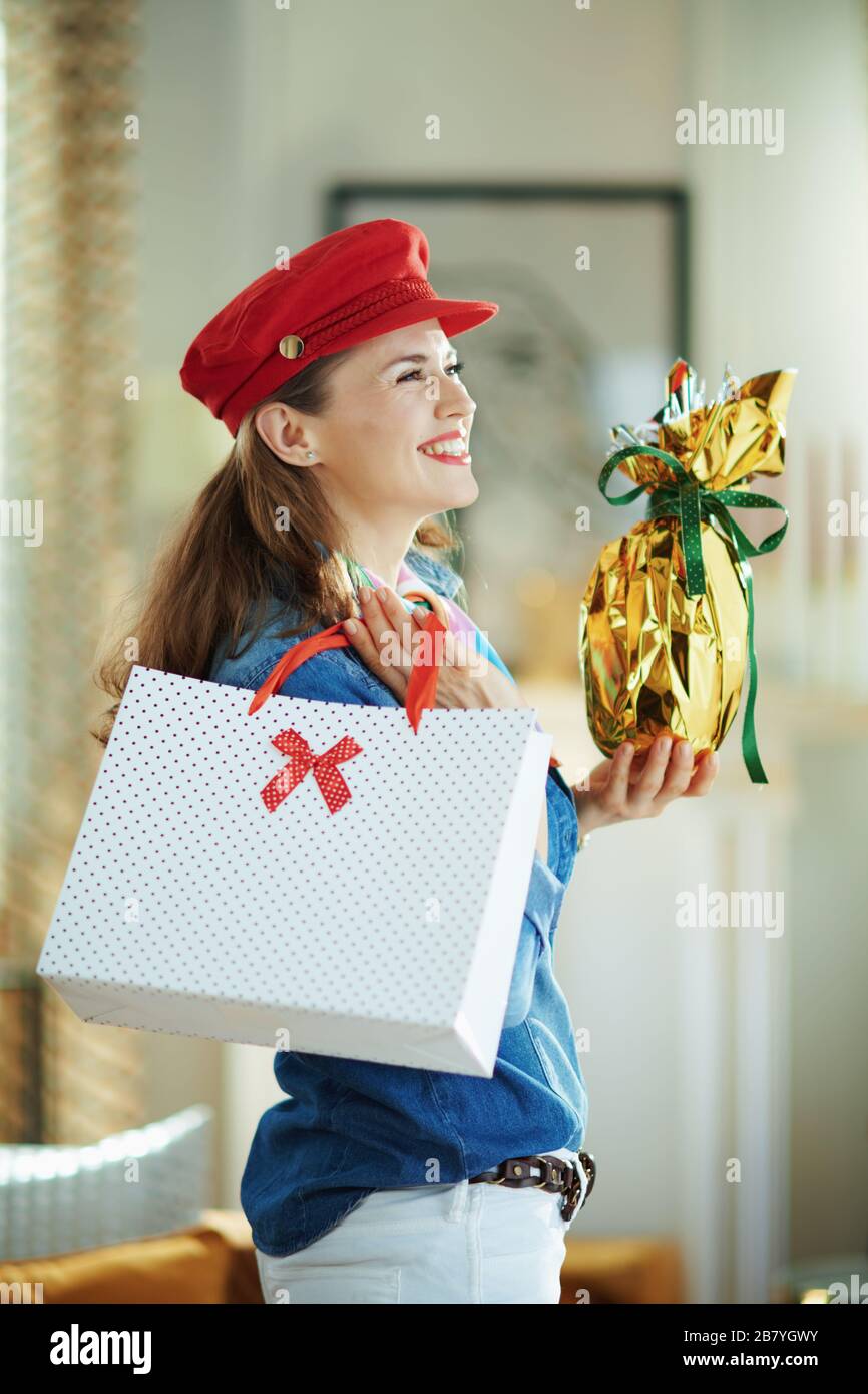 Seitenansicht einer lächelnden trendigen Frau in einem Jeans-Hemd und rotem Hut mit eingewickelter Goldfolie großes osterei und weißer Polka-Punkt-Einkaufstasche Stockfoto