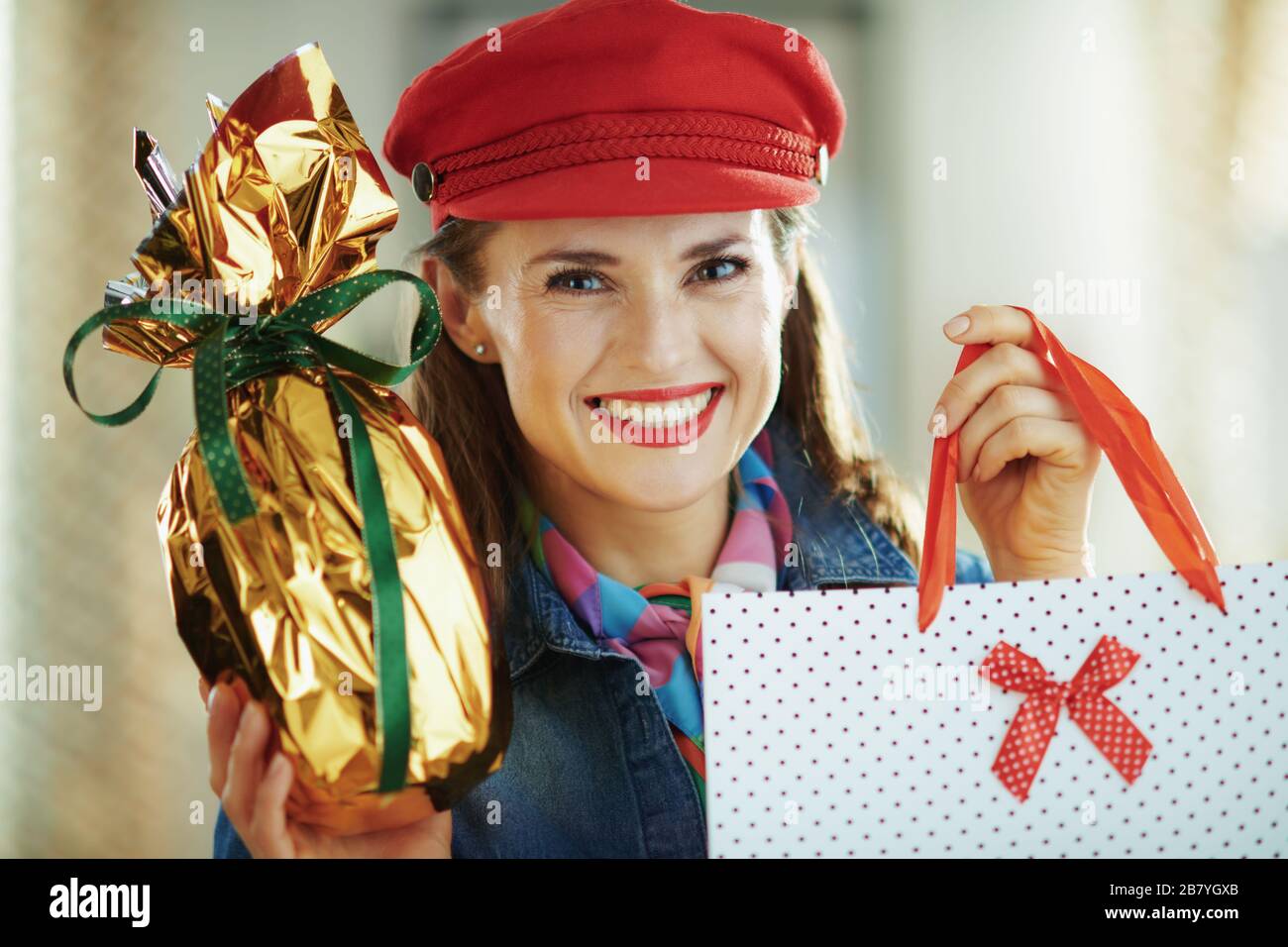 Porträt der fröhlichen jungen Frau in einem Jeans-Hemd und rotem Hut mit eingewickelt in Goldfolie großes osterei und weißer Polka-Punkt-Einkaufstasche im modernen Haus i Stockfoto
