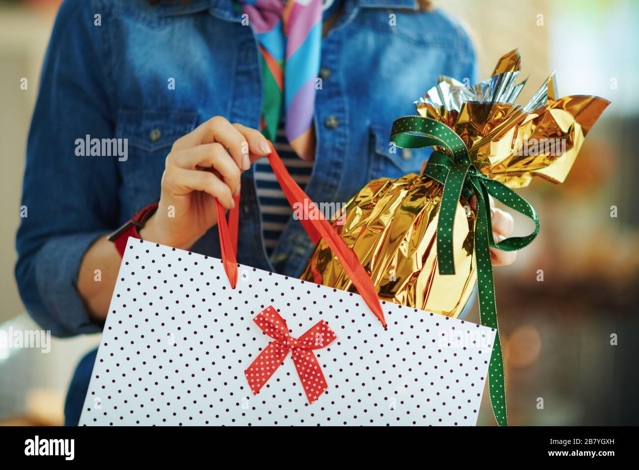 Nahaufnahme der Frau im Haus am sonnigen Frühlingstag, mit Goldfolie umhülltes großes osterei aus der weißen Einkaufstasche mit Punktmuster. Stockfoto