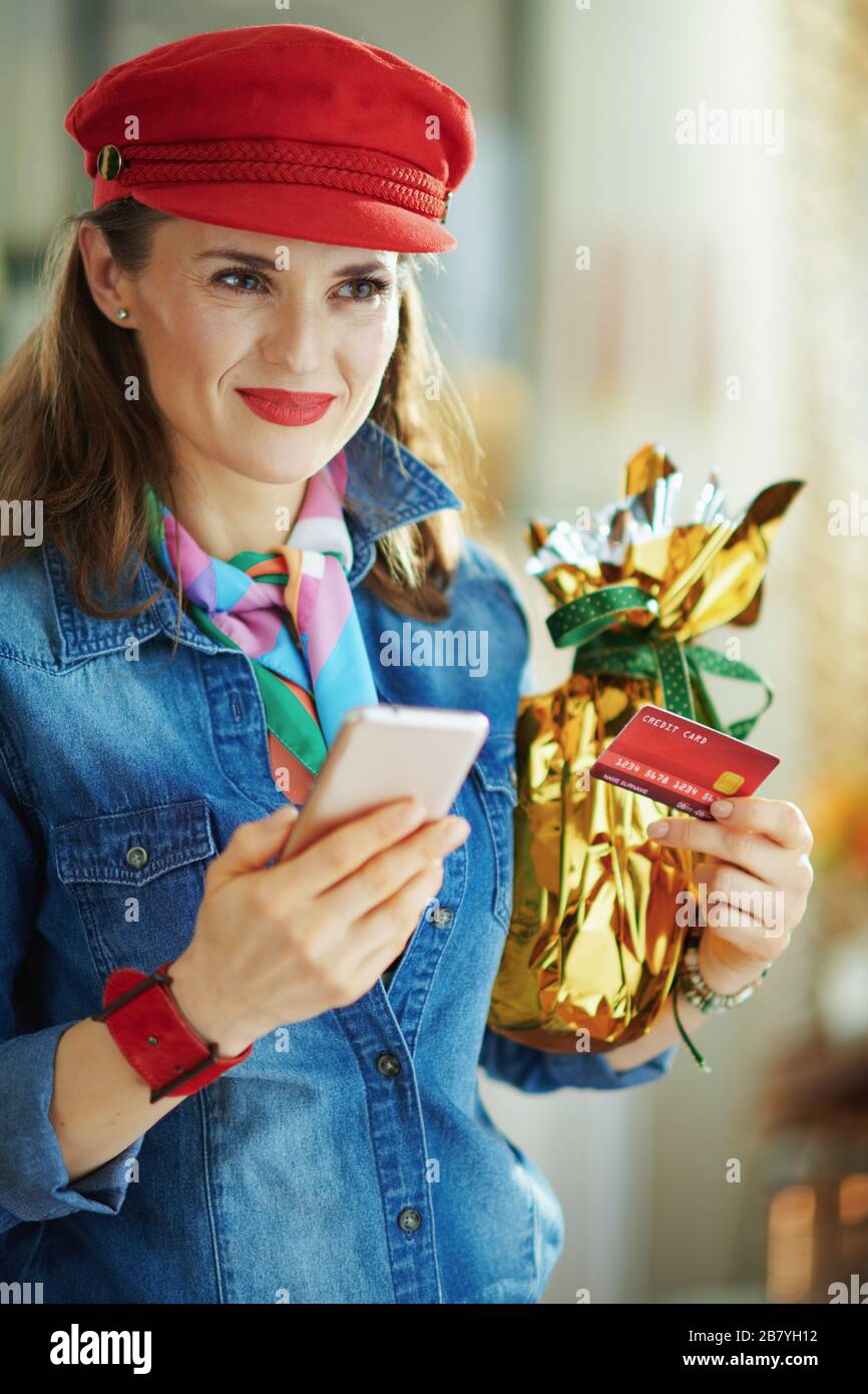 Junge junge Frau in Jeans-Hemd und rotem Hut mit Kreditkarte und in Goldfolie gehüllt großes osterei Online-Shopping auf der E-Commerce-Website machen Stockfoto