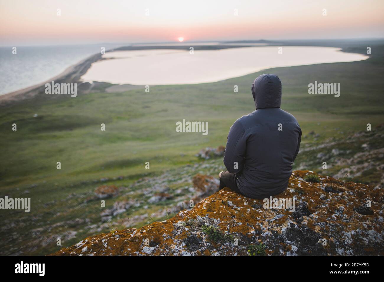 Junger Mann in Kapuzenjacke, der während des Sonnenuntergangs auf dem Hügel sitzt Stockfoto
