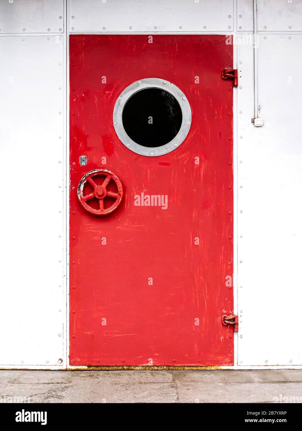 Rot lackierte Eisentür mit Porthole an weißer Fassadenwand Stockfoto