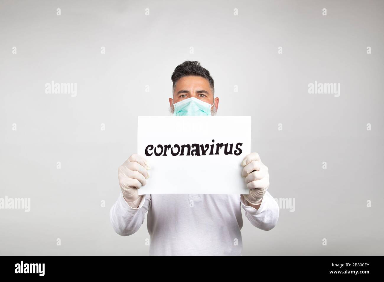 Porträt des Menschen in einer chirurgischen Maske, die ein weißes Schild mit dem Wort Coronavirus auf weißem Hintergrund hält. Stockfoto