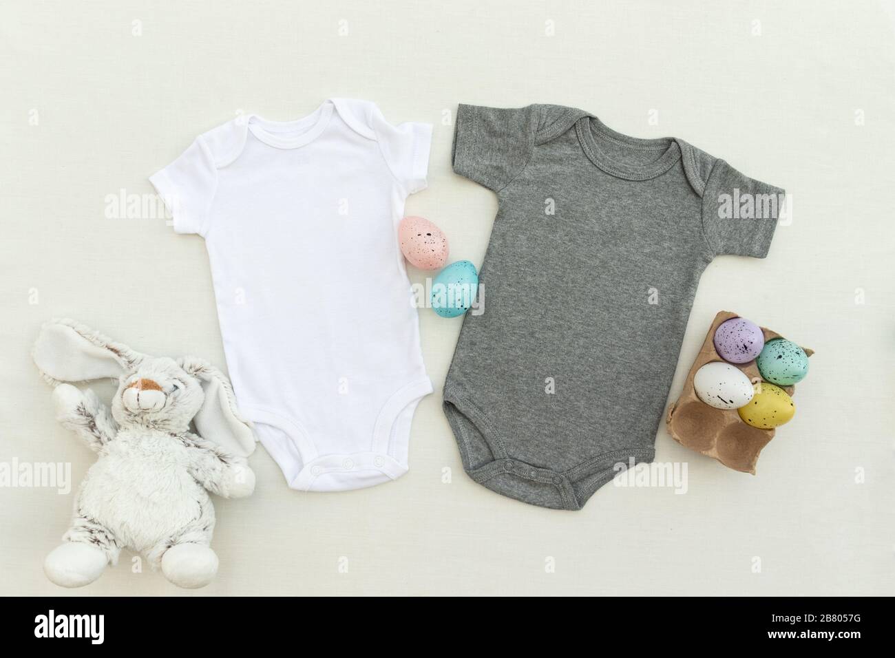 Zwei Oster-Baby-Bodys verspottet - zwei Osterbaby-Weste-Babys wachsen auf einem hellen Hintergrund mit Ostereiern und Hünenspielzeug Stockfoto