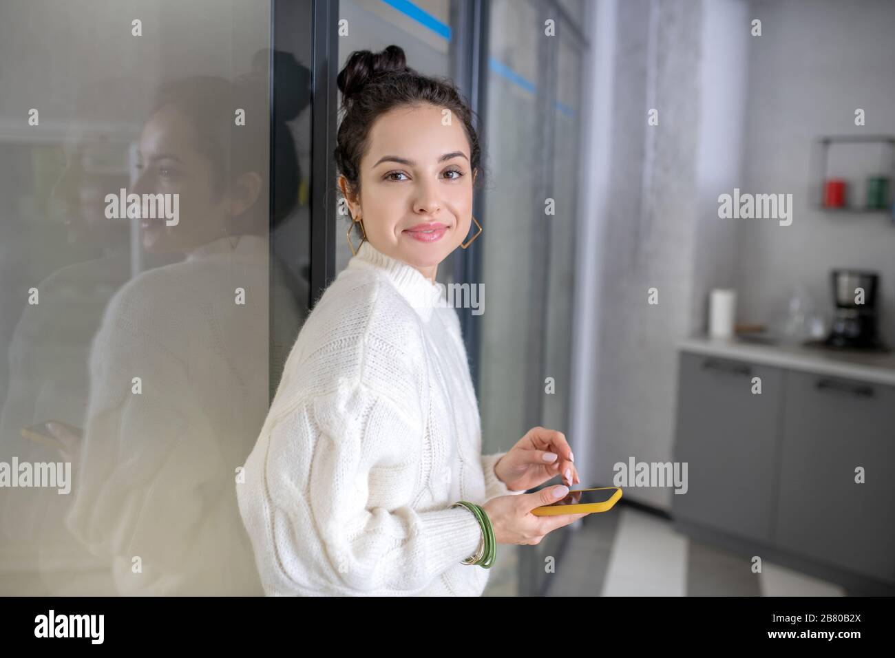 Junge Frau lehnt sich an die Glaswand, hält mobil und lächelt Stockfoto