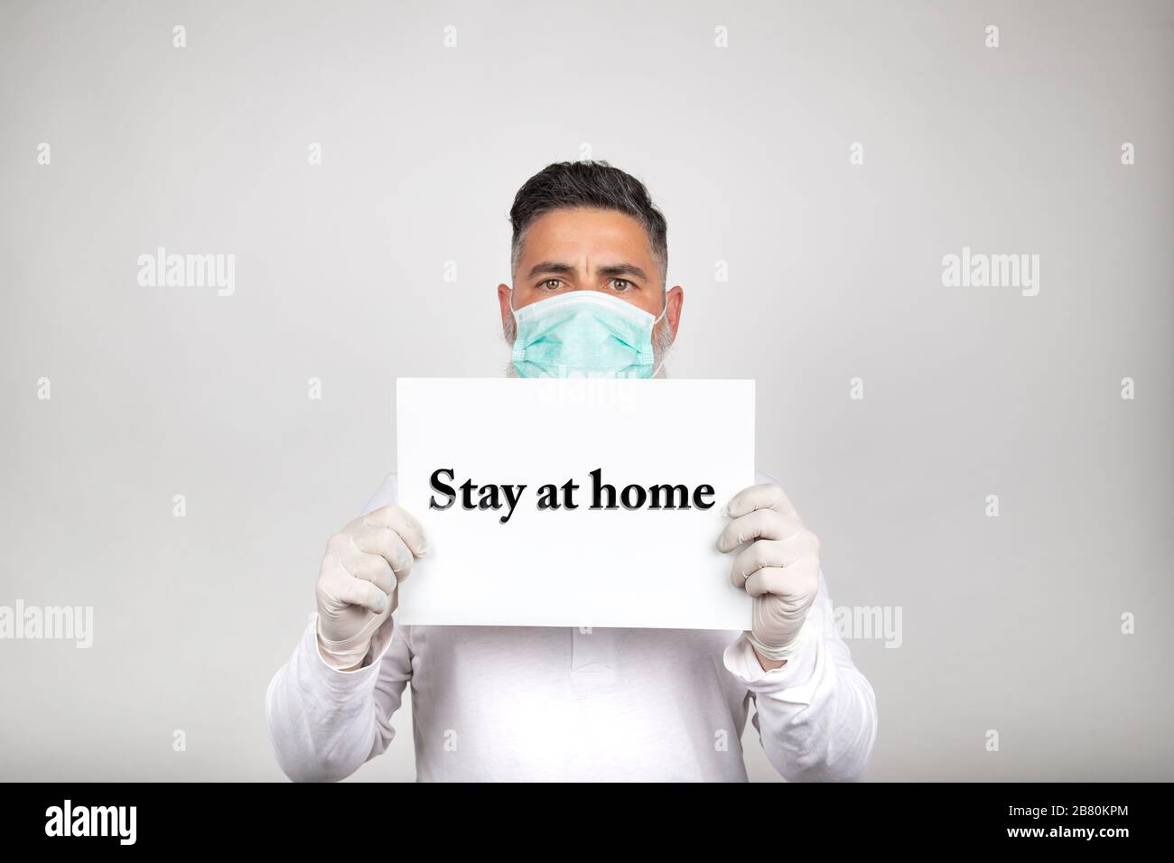 Porträt des Mannes in der chirurgischen Maske, das ein weißes Schild mit der Phrase hält, bleibt auf weißem Hintergrund zu Hause. Vorbeugung gegen Coronavirus Stockfoto
