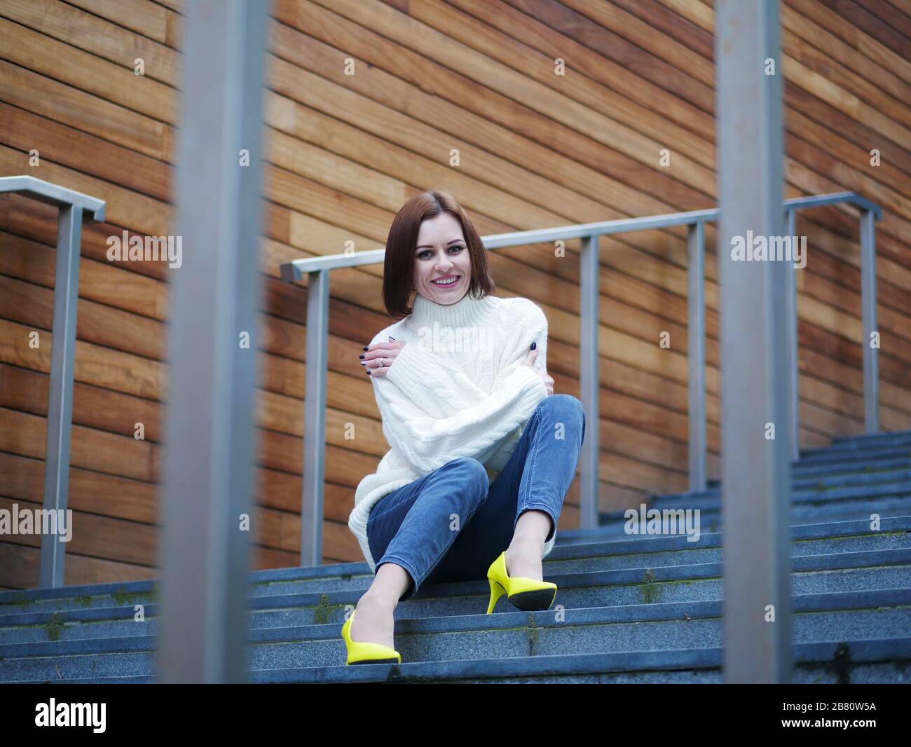 Bild einer schönen, stilvollen Frau in gelben Schuhen, die auf einer Straßentreppe mit gekreuzten Händen sitzt. Holzhintergrund. Stockfoto