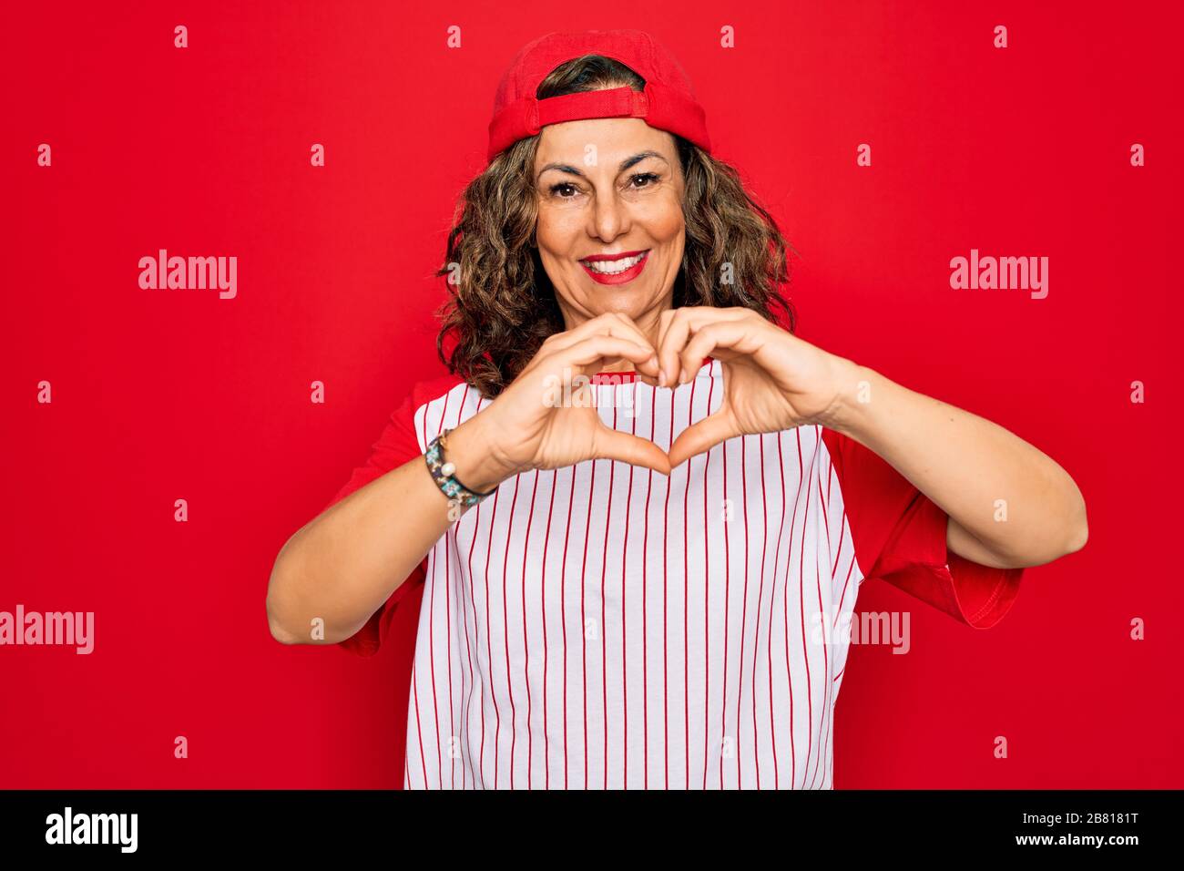 Seniorin im mittleren Alter, die Baseball-Equiment über rotem isoliertem Hintergrund trägt, lächelt in Liebe und zeigt Herzsymbol und Form mit den Händen. Romantische Kon Stockfoto