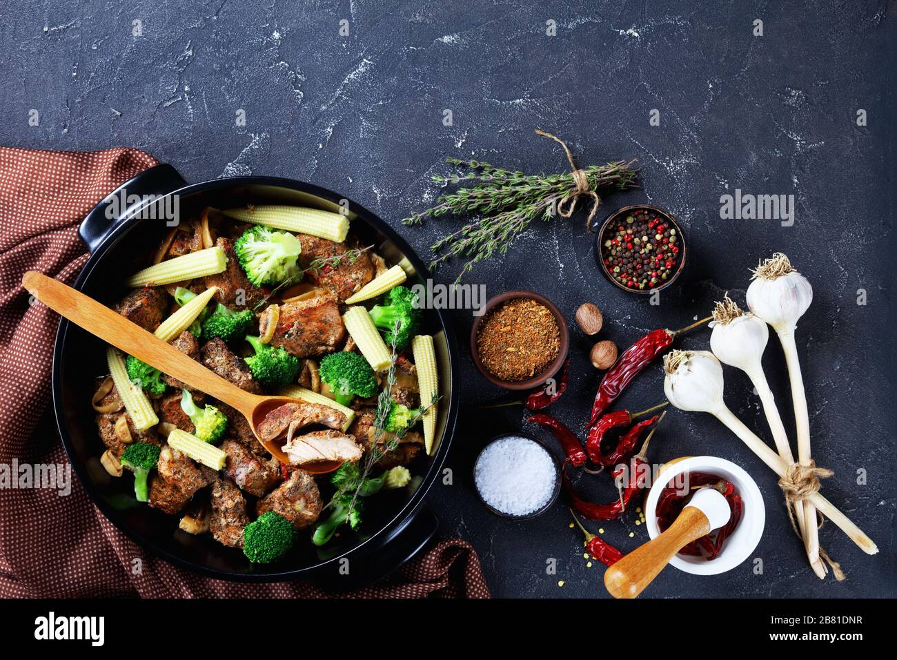 Schwarzes Gericht mit geröstetem Fleisch und Gemüse: Brokkoli, Mais, Zwiebel und Knoblauch mit Zutaten auf dunklem Betongrund: Pfeffer, Chili, Thymian, Stockfoto