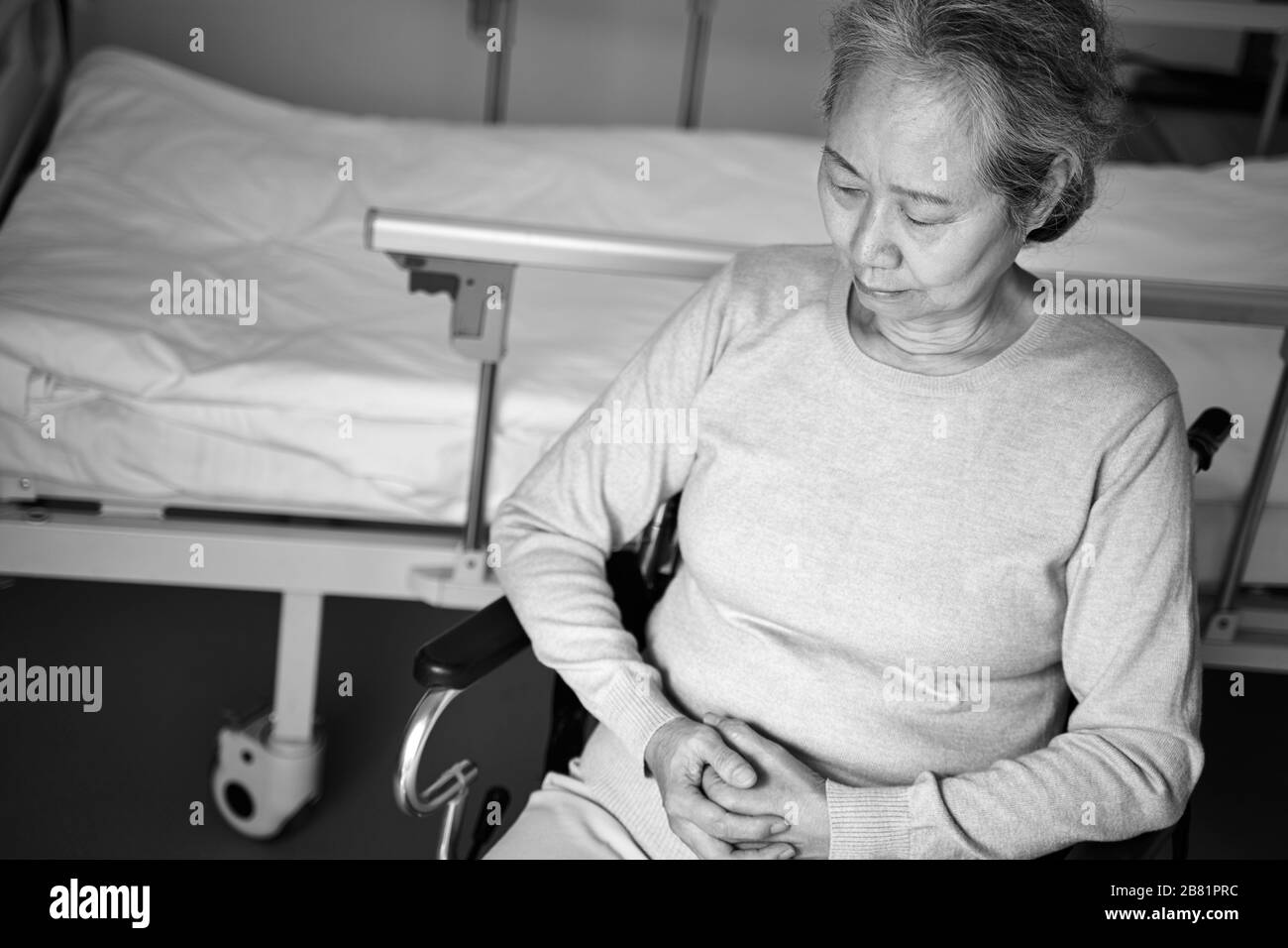 Hochwinkeliger Blick auf die traurige asiatische Frau, die im Rollstuhl neben dem Krankenhausbett sitzt, schwarz-weiß Stockfoto
