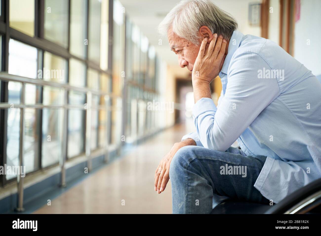 Trauriger und verwüsteter asiatischer Obermann, der allein in einem leeren Krankenhausflur sitzt, Kopf in der Hand Stockfoto