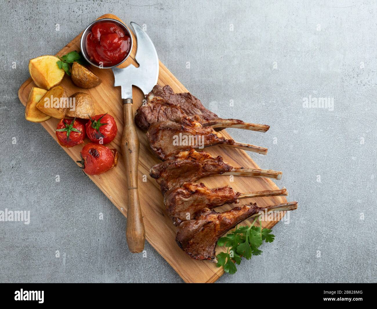 Gebratene Lammkoteletts auf einem Holzbrett mit Kartoffeln und Tomaten aus roter Soße Stockfoto