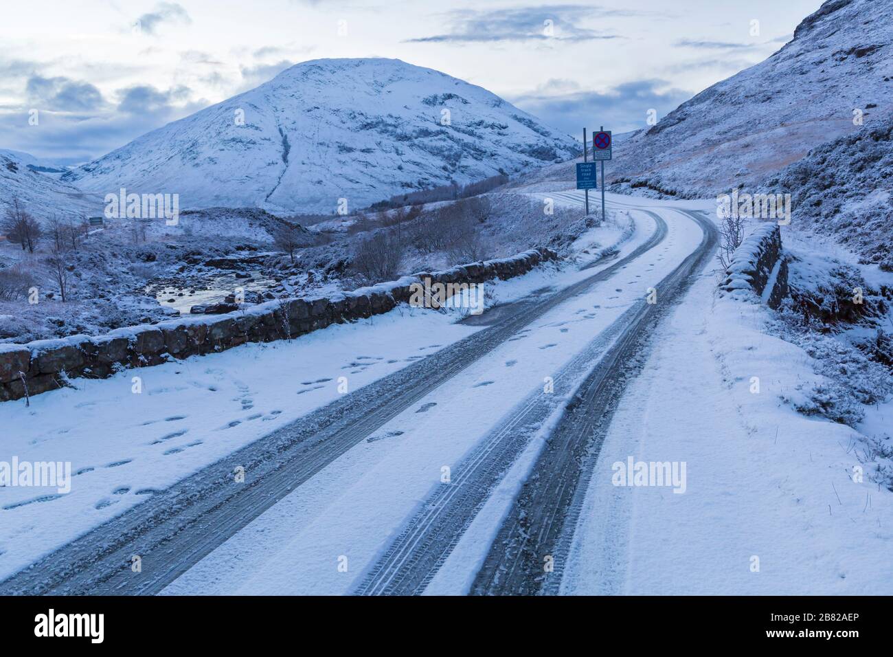 Reifenspuren und Fußabdrücke auf schneebedeckten Straßen, die im Winter in Glencoe, Highlands, Schottland durch die Berge biegen Stockfoto