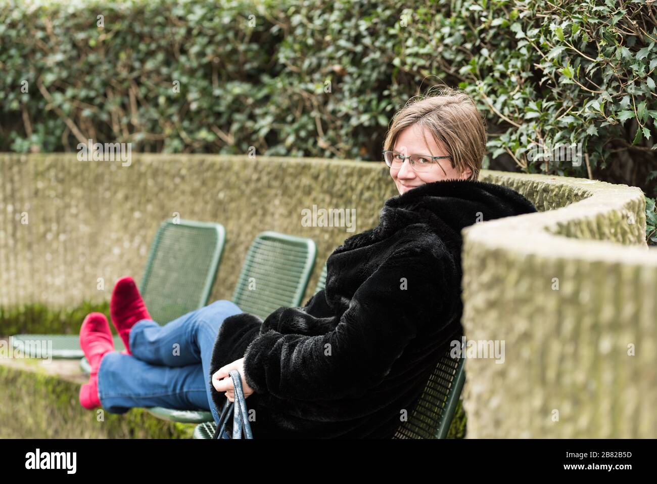 Attraktive dreißigjährige Frau sitzt auf einer gebogenen Bank mit roten Schuhen Stockfoto