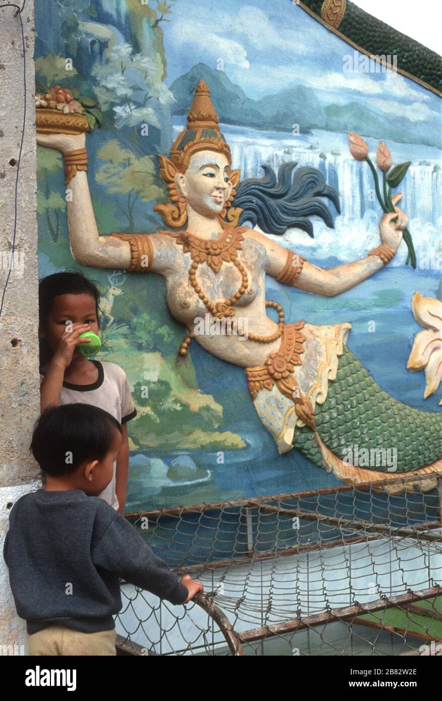 Zwei junge Lao-Jungen ruhen und einer genießt ein Getränk neben einem Wandbild einer Meerjungfrau in Pha that Luang (The Great Stupa) in Vientiane, Laos. Sie hält lotosblüten und eine Schüssel/einen Obstkorb. Sie trägt eine traditionelle Kopfbeere und eine Kette von Perlen. Das Bild zeigt Berge, einen Fluss und einen Wasserfall im Hintergrund. Stockfoto