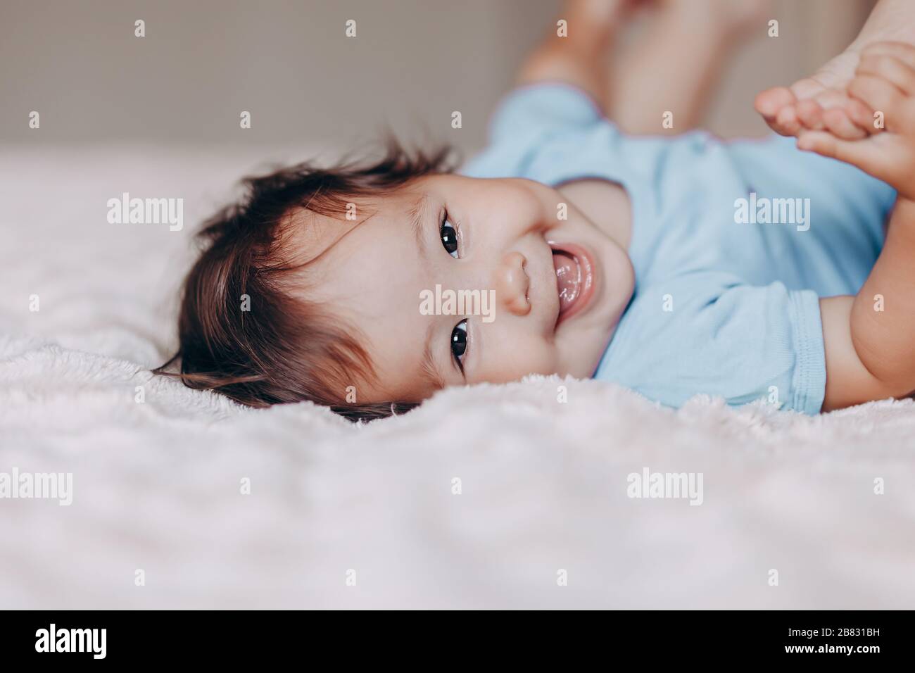 Süßes Lachen ein einjähriges Mädchen, das auf dem Bett liegt und die Kamera betrachtet, berührt ihre Füße Stockfoto