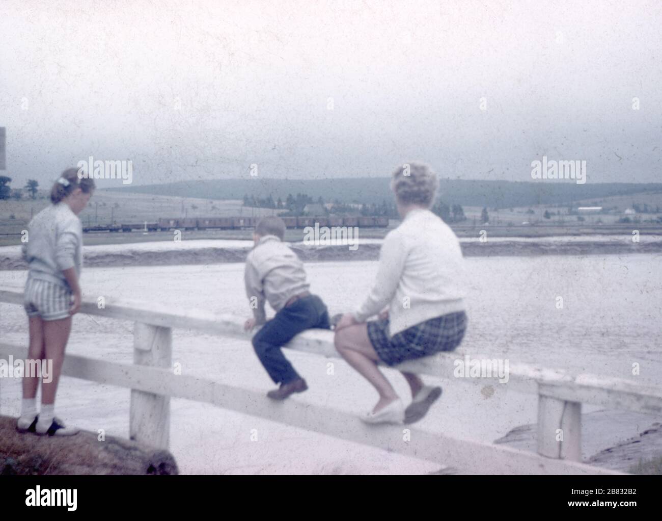 Eine Mutter und ihre beiden Kinder balancieren an einem weißen Zaun über einer Klippe, die zu einem Gewässer unterhalb führt, an einem übergiebelten Tag, 1960. () Stockfoto