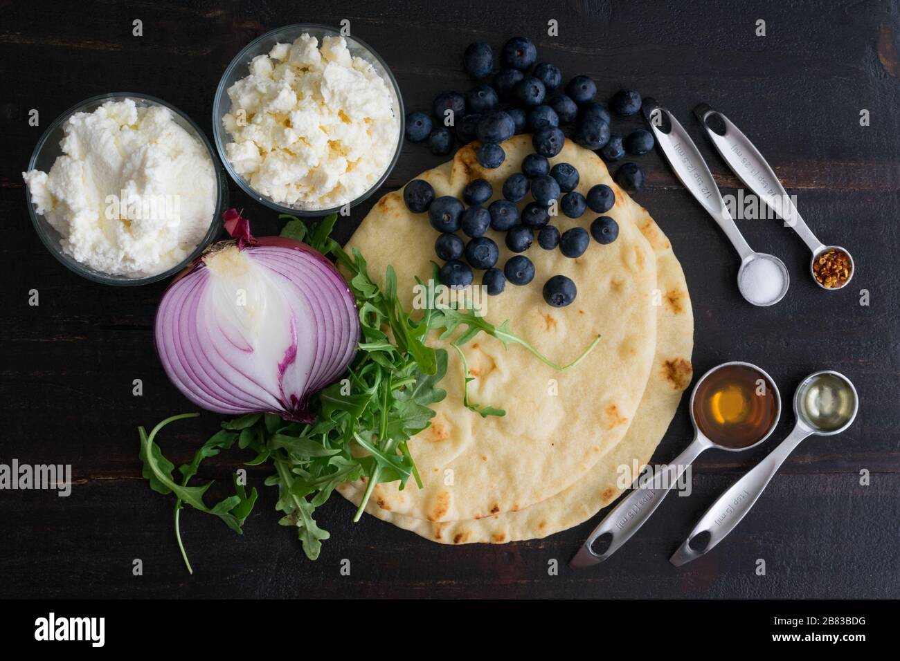 Zutaten für Blueberry, Feta und Honeykaramelisierte Zwiebel Naan Pizza: Naanische Fladenbrote, Arugula und andere Zutaten auf einem Holztisch Stockfoto