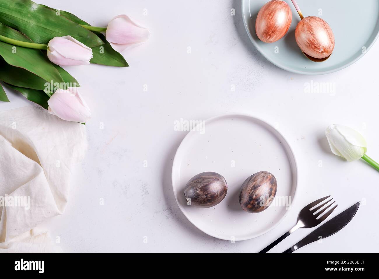 Grußkarte für Ostern mit handgemalten, hellen Eiern auf Tellern und Tulpenblumen auf hellgrauem Marmorhintergrund. Stockfoto