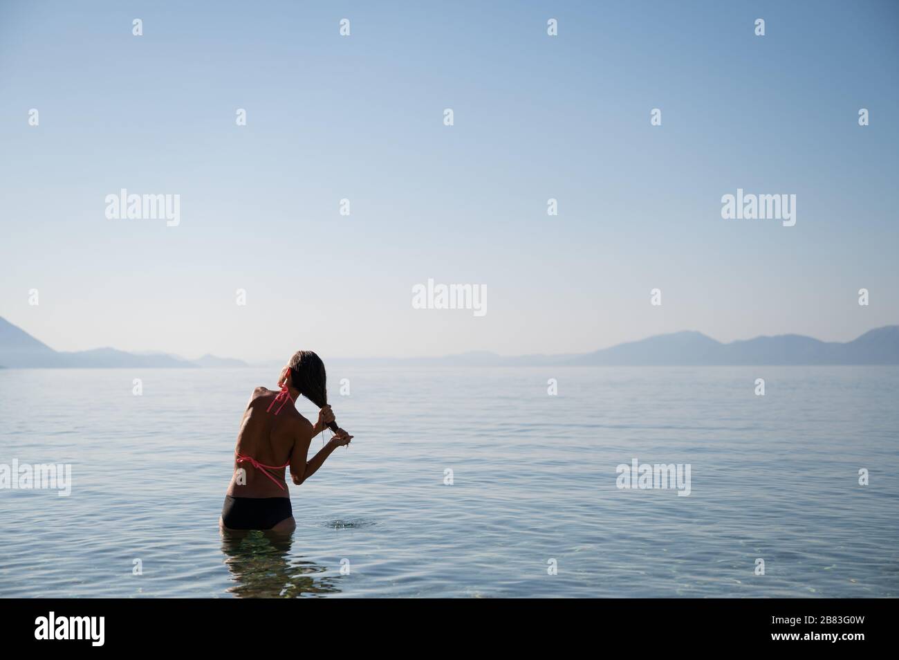 Blick von hinten auf eine junge Frau, die nach einem Schwimmen in ruhiger Morgenmeere steht und ihr nasses Haar zusammendrückt. Stockfoto
