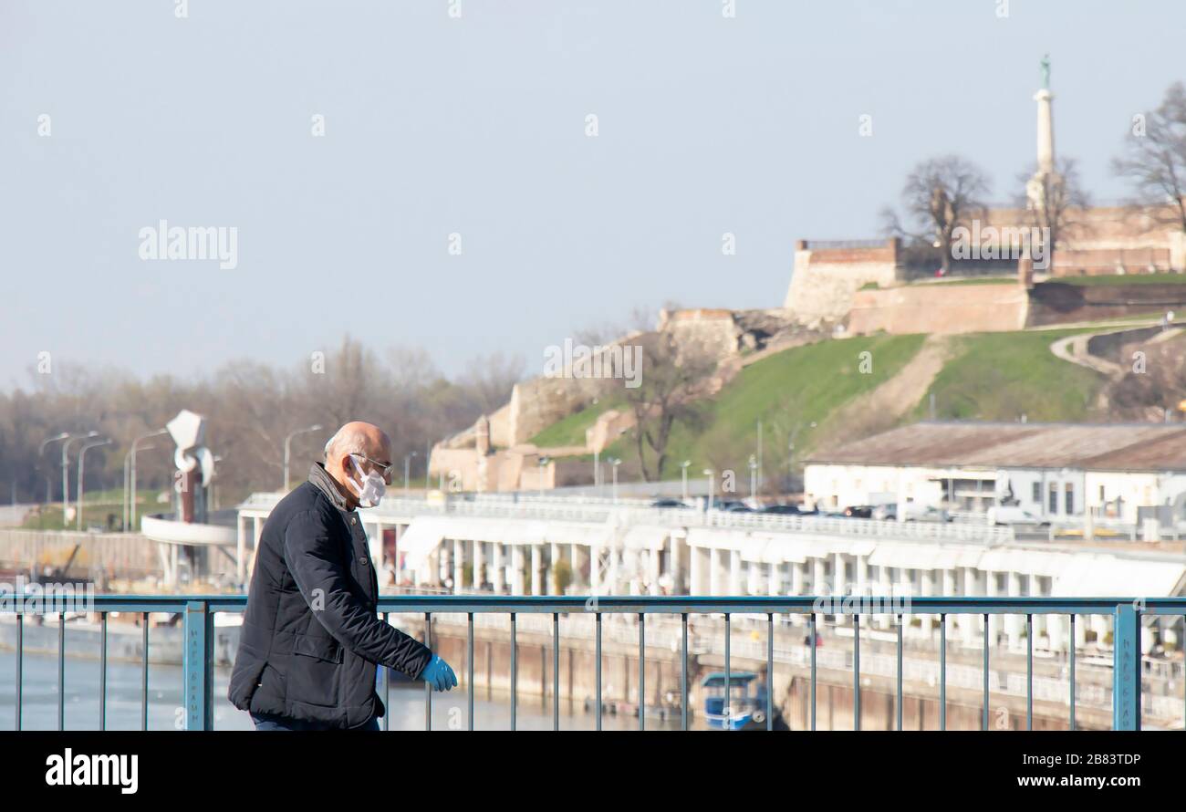 Belgrad, Serbien - 18. März 2020: Ältere Männer, die Schutzmasken tragen, die allein auf der Stadtbrücke mit der Festung Kalemegdan im Hintergrund in der ti spazieren gehen Stockfoto