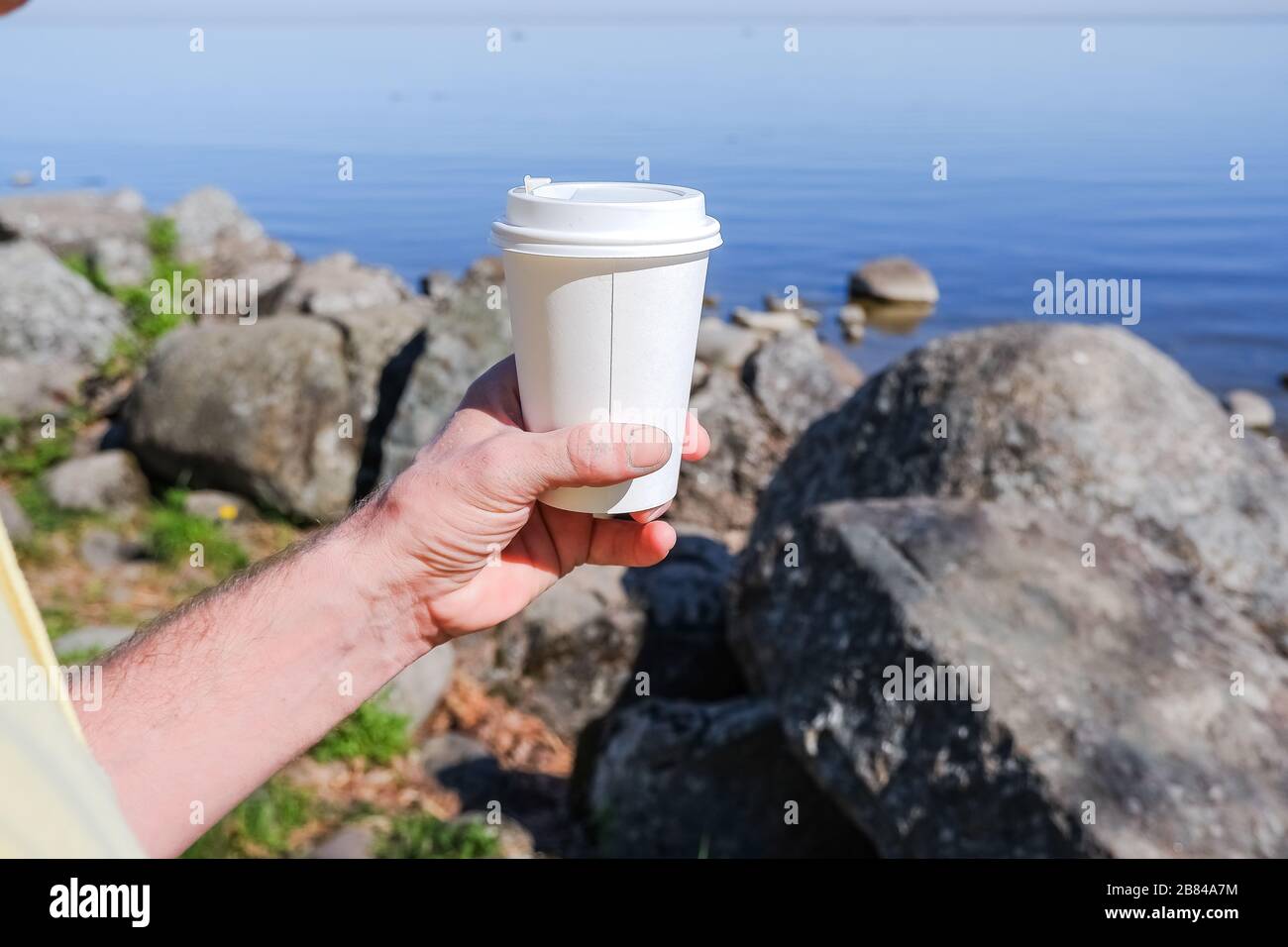 Morgen Papierkaffeetasse mit leckeren heißen Kaffee bei Männern Hand im Freien auf Meeresgrund.Hand hält ein weißes Papppapier Becher für Getränke zum Mitnehmen Stockfoto