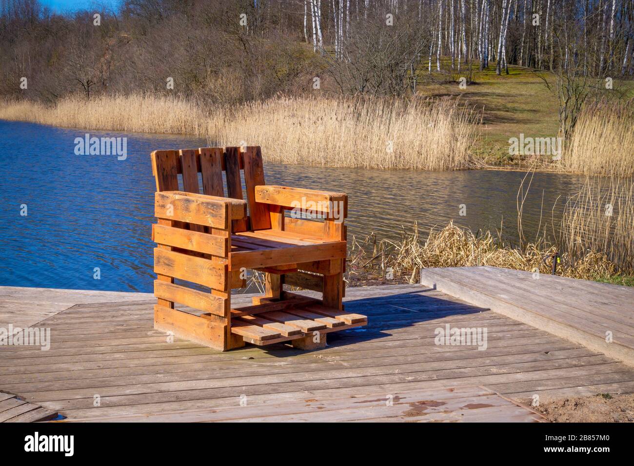 Rustikaler handgefertigter Stuhl aus Holzpaletten auf Holzdeck mit Blick auf einen Fluss oder See mit Schilf bei Frühlingssonne Stockfoto