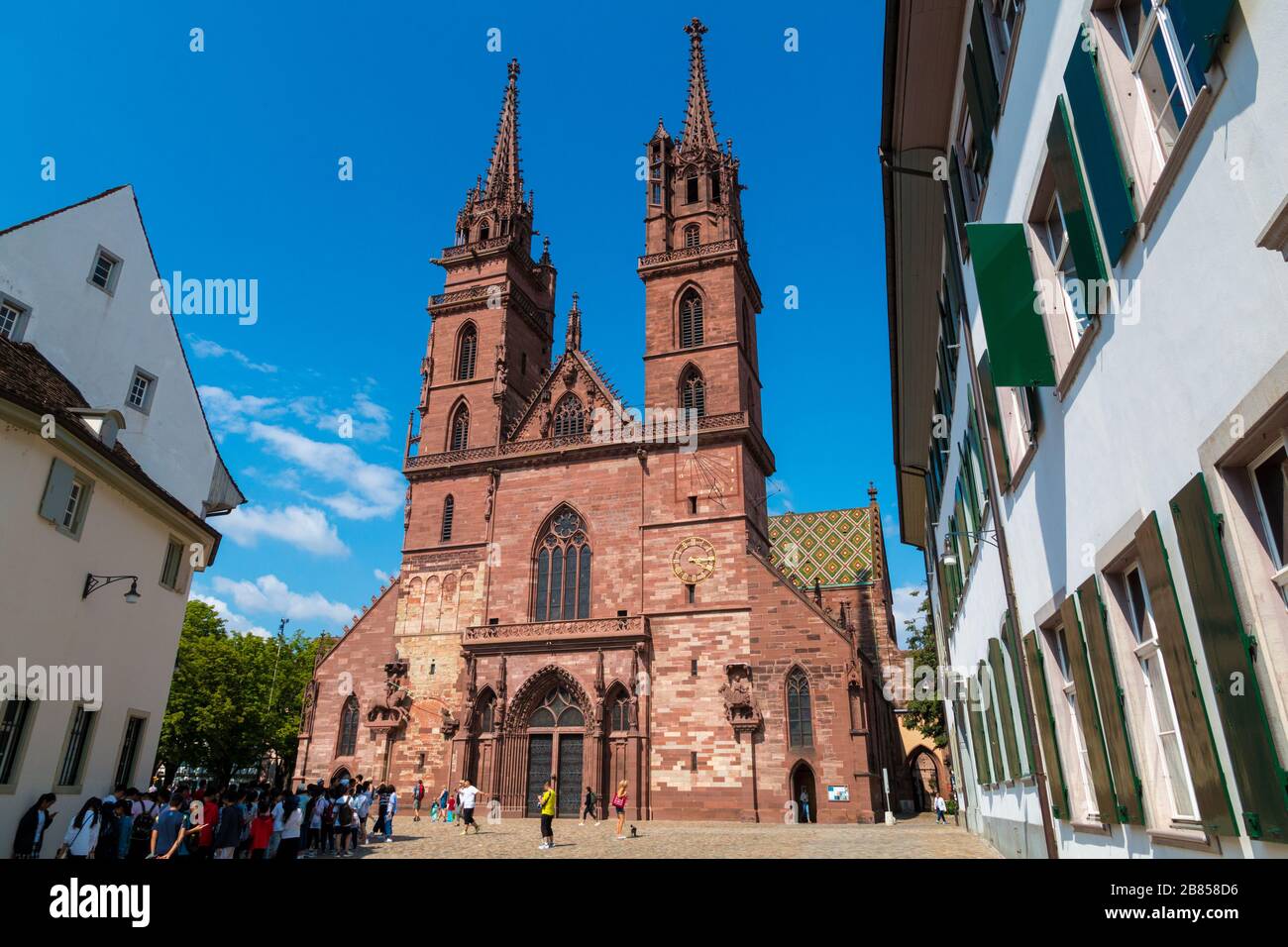 Eine Gruppe von Touristen wartet vor dem Basel Minster an einem schönen sonnigen Tag mit blauem Himmel. Mit seinen roten Sandstein-Wänden, bunten Dachziegeln... Stockfoto