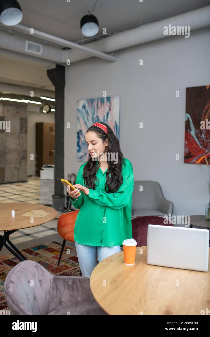 Hübsche Frau, die das gelbe Smartphone in den Händen, im Büro, betrachtet. Stockfoto