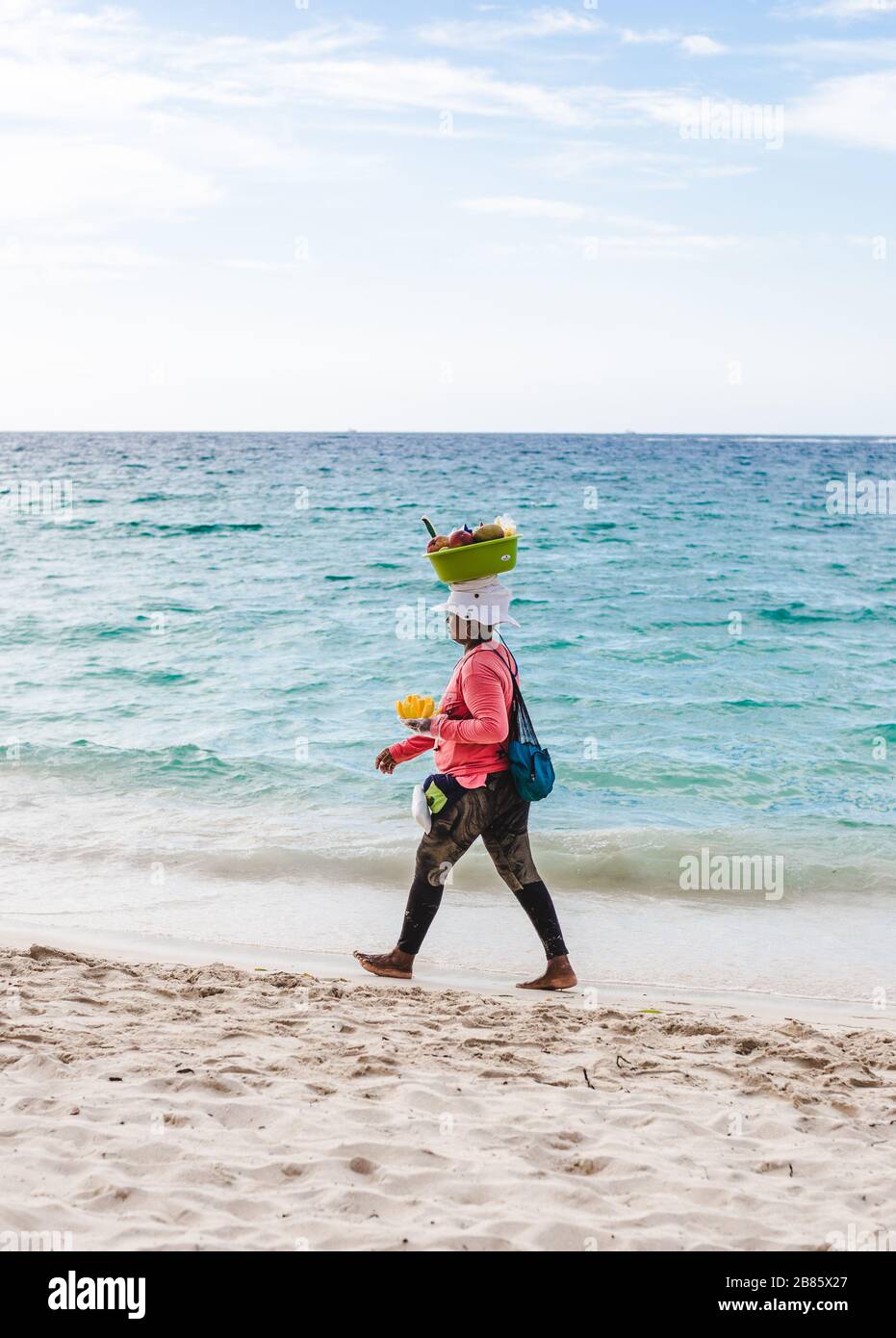 Frau aus der Region spaziert am weissgeschliffenen Strand von Isla Baru hinunter, um Obst aus einem Korb auf dem Kopf in der Nähe von Cartagena, Kolumbien zu verkaufen Stockfoto