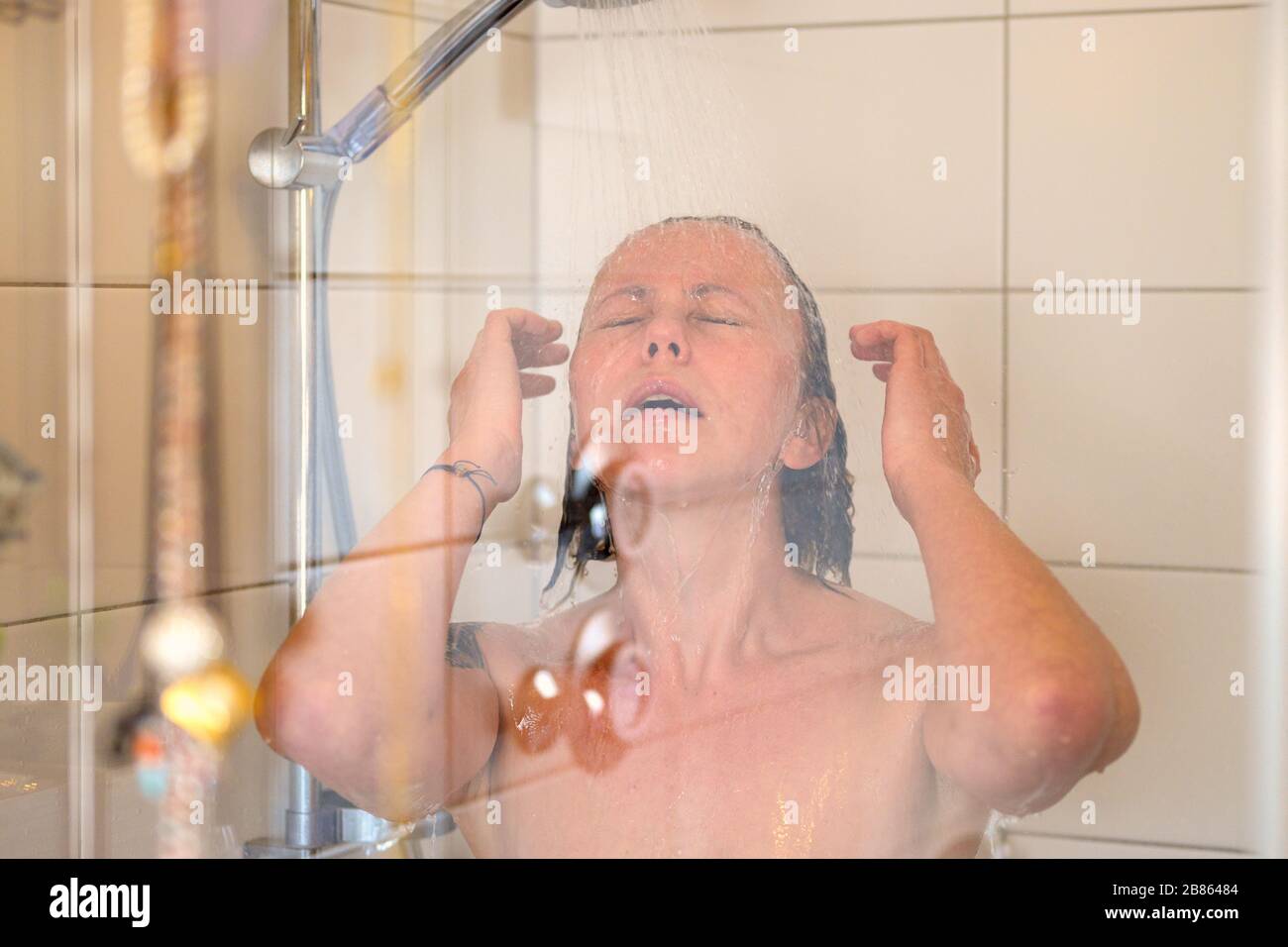 Frau, die eine dampfende heiße Dusche genießt, die ihr Haar unter fließendem Wasser mit geschlossenen Augen in einem Nahporträt durch die Glaskabine in abspült Stockfoto