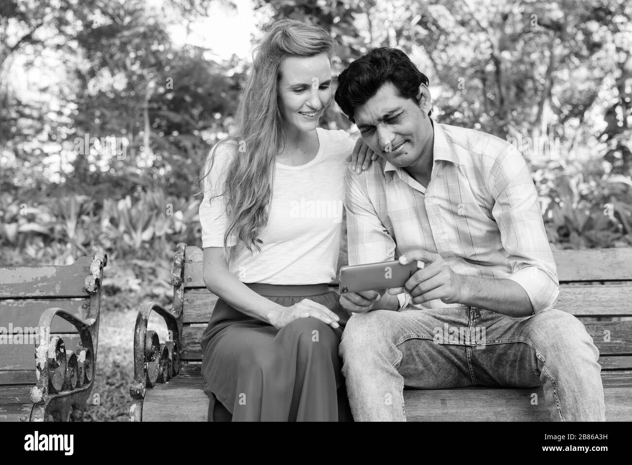 Fröhliches multiethnisches Paar lächelt, während es mit dem Handy telefoniert und zusammen auf einer Holzbank sitzt, die sich in den Park verliebt Stockfoto