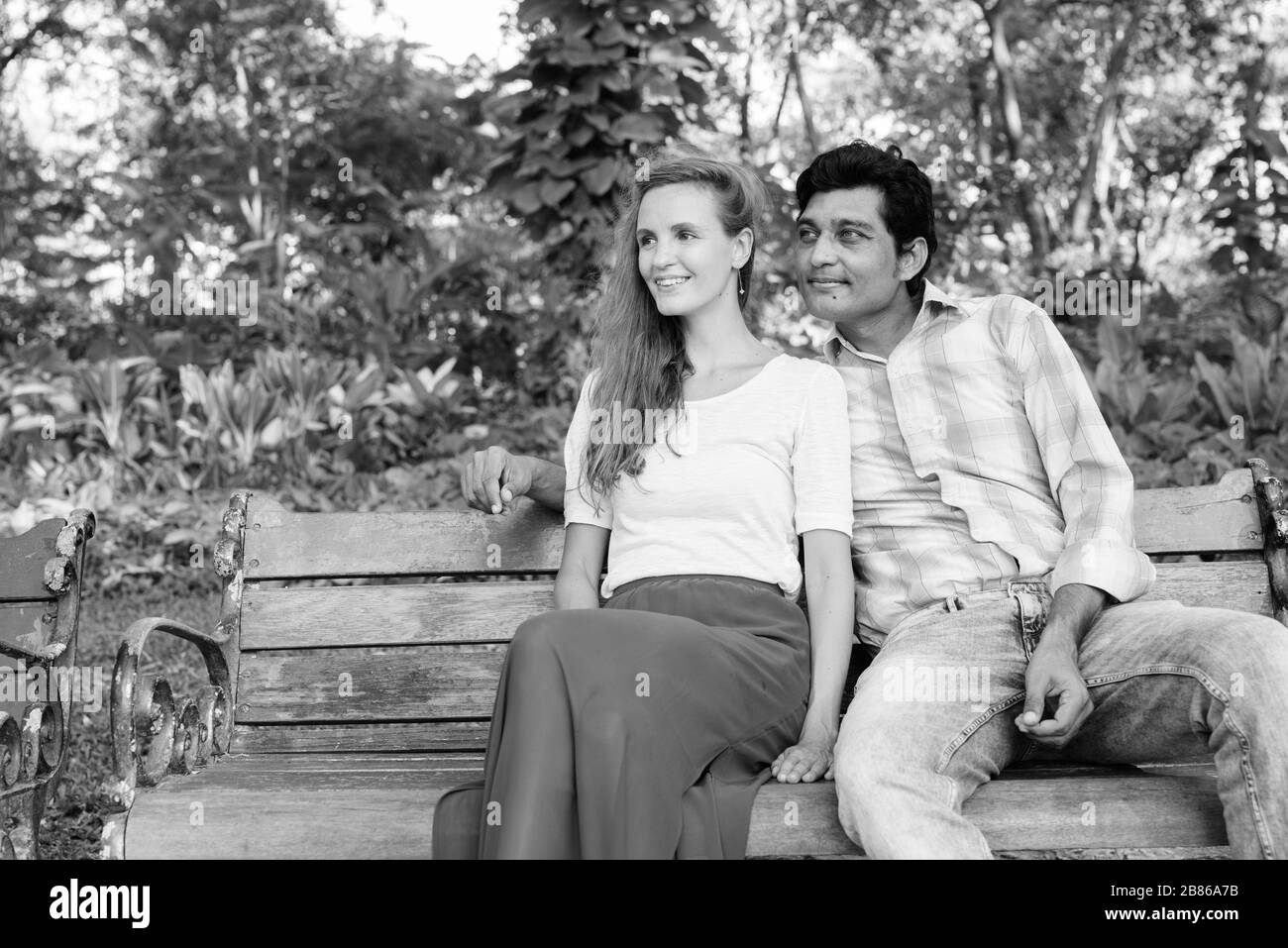 Fröhliches multiethnisches Paar lächelt und denkt, während es auf einer Holzbank im Park sitzt Stockfoto