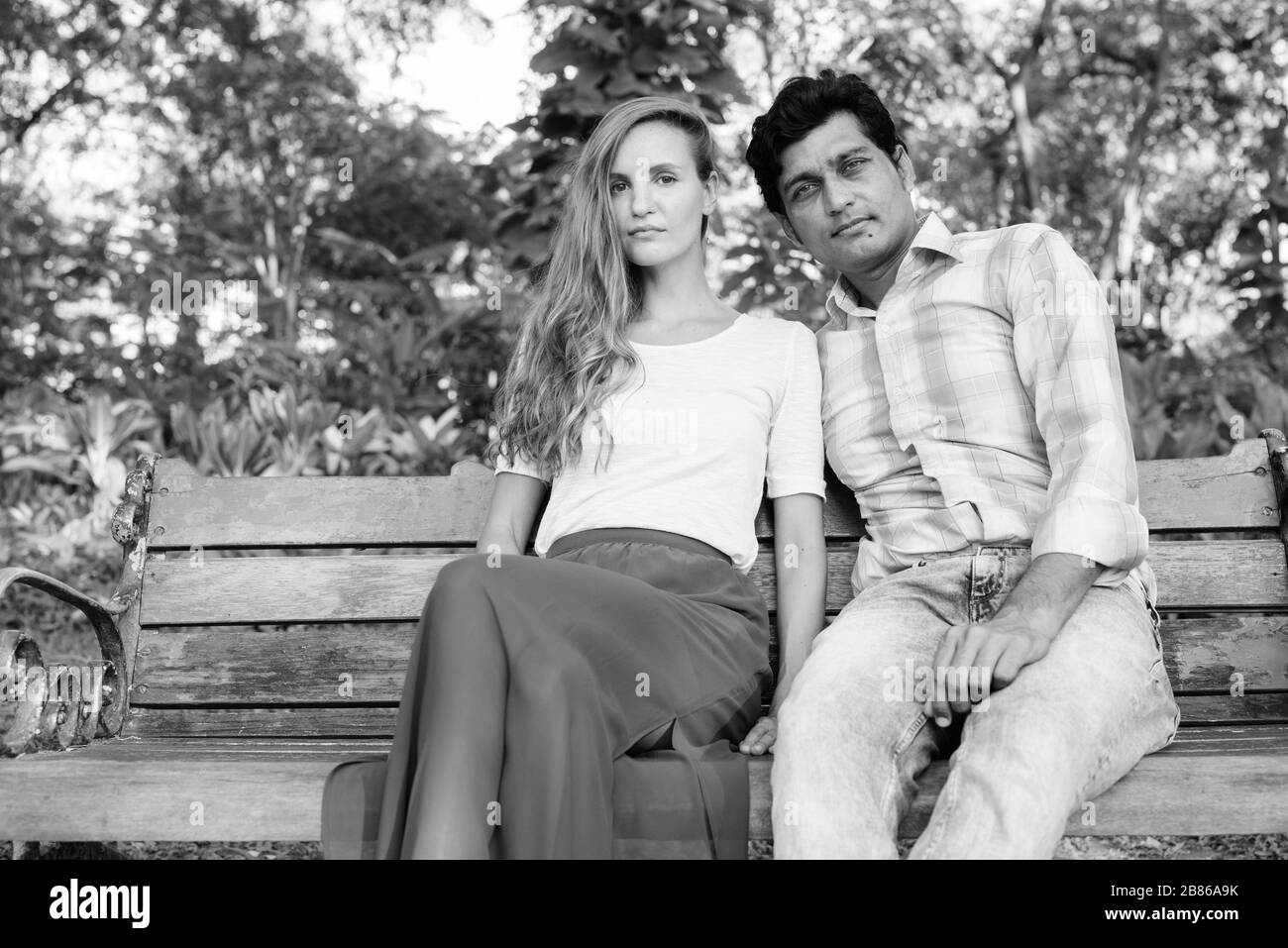 Ein Paar aus verschiedenen ethnischen Gruppen, das auf einer Holzbank sitzt und sich in den Park verliebt Stockfoto