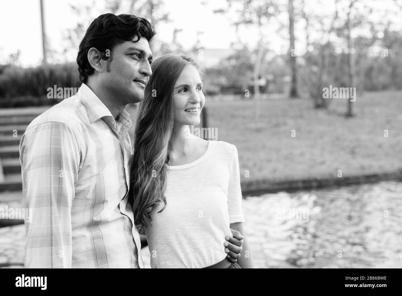 Fröhliches multiethnisches Paar lächelt, während man auf dem Steinweg mitten im See gegen den Blick auf den Park denkt Stockfoto