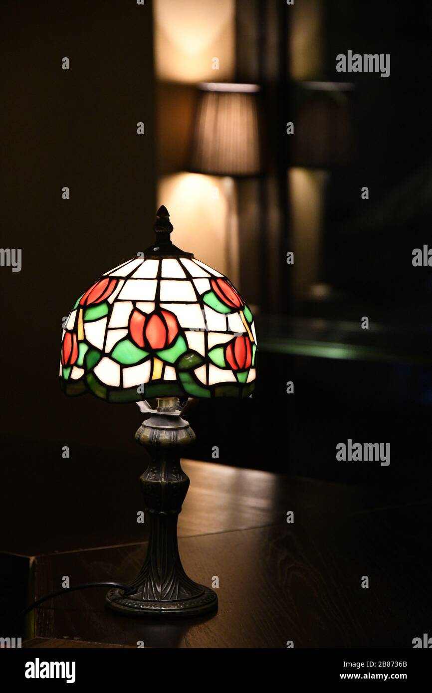 Lampenschirm aus Buntglas mit Blumenmuster. Glühende Mosaiklampe im Nahaufnahme auf dunklem, verschwommem Hintergrund. Vintage-Tischleuchte mit verzierten Lampenschatten Stockfoto
