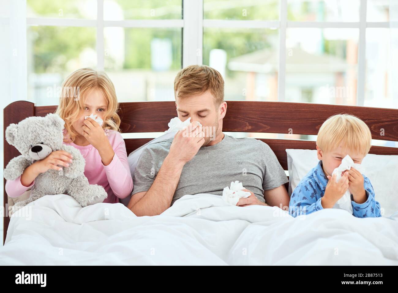 Der junge Vater und seine Kinder leiden an Grippe oder Kälte und wischen ihre Nasen, während sie zusammen im Bett liegen. Viruserkrankung. Coronavirus Konzept. Kranke Familie zu Hause. Gesundheitskonzept Stockfoto