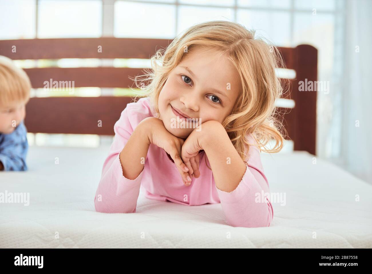 Portrait eines niedlichen kleinen Mädchens in pinkfarbenem Pajama, das mit der Kamera lächelt, während sie mit ihrem Bruder auf einer großen und weichen weißen bequemen Matratze liegt. Quarantäne. Schlafzimmer. Familie Stockfoto
