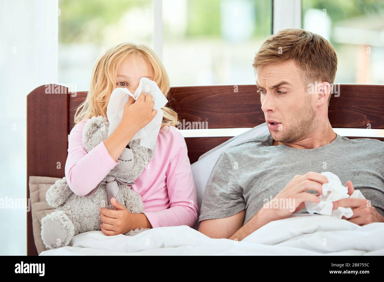 Junger Vater und Tochter, die an Grippe oder Kälte erkrankt sind, haben Runennasen, während sie zuhause im Bett zusammen ruhen. Viruserkrankung. Coronavirus Konzept. Kranke Familie zu Hause. Gesundheitskonzept Stockfoto