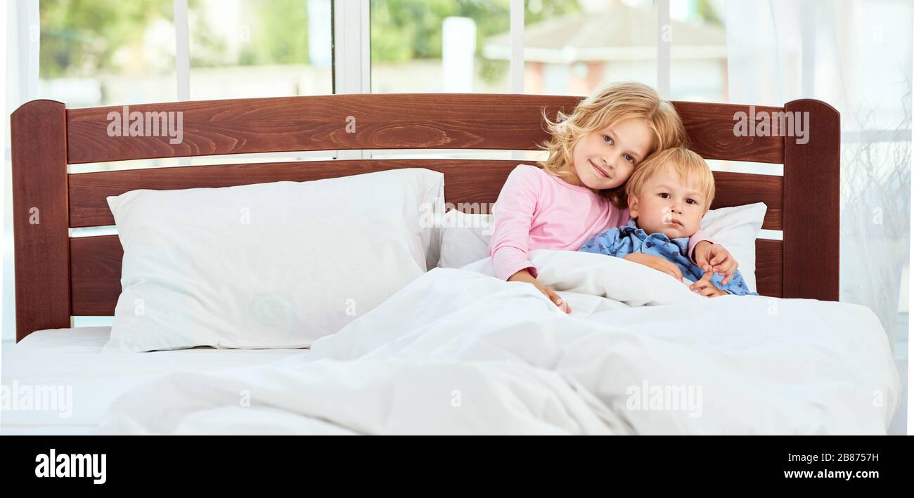 Süßer kleiner Bruder und Schwester im Schlafanzug und Blick auf die Kamera mit Lächeln, während sie zusammen in einem großen Bett zu Hause liegen. Quarantäne. Genießen Sie das Wochenende zu Hause. Familie Stockfoto