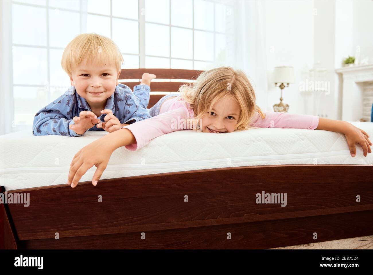Fröhlicher, süßer kleiner Bruder und Schwester, die die Kamera angucken und lächeln, während sie auf einer großen, bequemen weißen Matratze im Schlafzimmer liegen. Gemeinsam spielen. Quarantäne. Schlafzimmer. Familie Stockfoto
