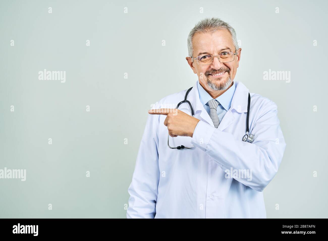 Fröhliche reife Arzt posing und lächelnd an Kamera, Gesundheitswesen und Medizin. Auf blauem Hintergrund isolieren. Hand bei copy Raum zeigt. Stockfoto