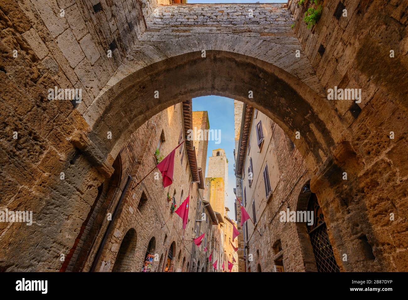 Ein Bogen über der Straße Via S. Matteo mit roten Flaggen, die bei Sonnenuntergang an den Wänden im historischen Zentrum von San Gimignano, Provinz Siena, Italien, auffliegen. Stockfoto