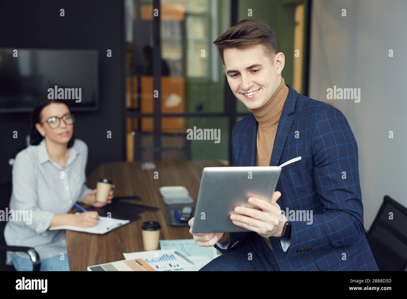 Junger Geschäftsmann, der Tablet-pc hält und ihn für die Online-Kommunikation mit Geschäftsfrau verwendet, die im Hintergrund im Büro sitzt Stockfoto
