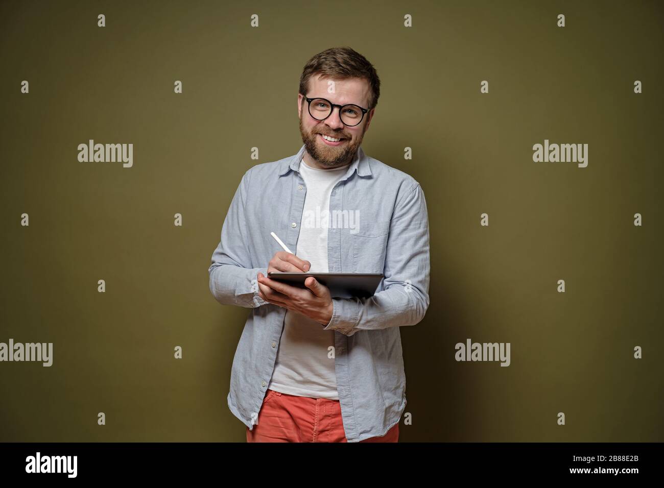 Der freundliche, bärtige Mann in der Brille arbeitet mit einem Tablet- und Stylus Stift, lächelt und blickt auf die Kamera, auf einem grünen Hintergrund. Stockfoto