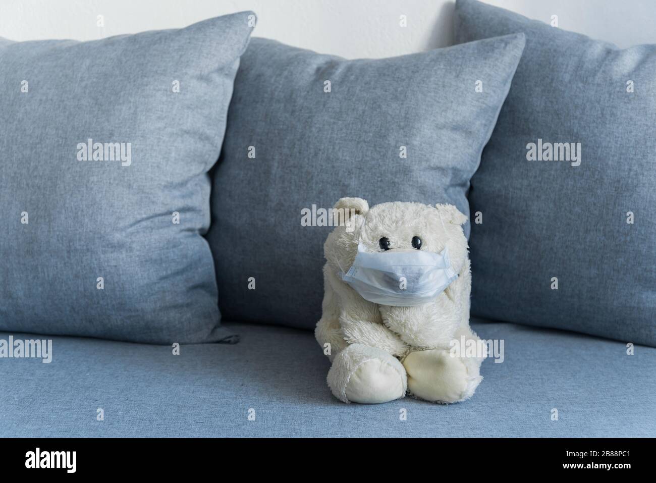 Weißer Teddybär sitzt in Selbstisolation auf grauem Sofa und trägt eine medizinische Maske Stockfoto